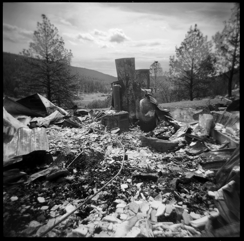 Black and white film Holga plastic camera photograph of a burned and charred black rubble of a house with a burned stove and well pump in the middle.
